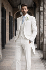 Man wearing a white morning suit with waistcoat standing in a classic stone corridor.